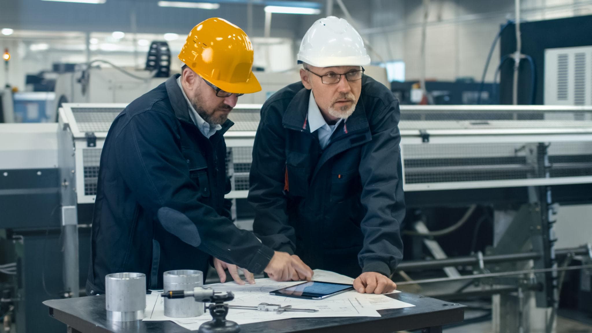 Twee technische professionals bekijken tekeningen en een tablet in een werkplaats